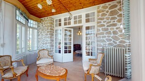 Ein Zimmer mit Steinwänden, Holzboden und großen Fenstern. Es gibt zwei Polsterstühle und einen runden Tisch.
