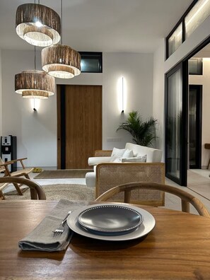 Un comedor con mesa de madera, un plato, una servilleta doblada y un tenedor. Hay sillas de mimbre, un sofá y una planta en maceta.