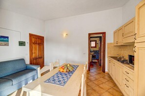 A dining area with a table set for two, a blue sofa, and a kitchen with wooden cabinets and a tiled backsplash. - Chic Sardinian Escape by the Beach (Badesi)