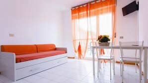A room with a white sofa, a dining table with chairs, a TV mounted on the wall, and a window with sheer curtains.