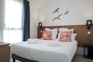 Ein ordentlich bezogenes Bett mit weißen Leinentextilien und Zierkissen, ein hölzernes Kopfteil, Wandlampen und ein Fenster mit Vorhängen.