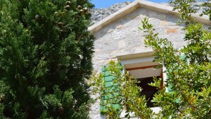 Un edificio in pietra con tetto spiovente, una finestra con persiane verdi e un albero in primo piano.