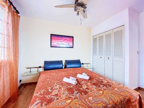 Une chambre d’hôtel avec un lit, deux chaises bleues, une petite table, un ventilateur de plafond, un tableau et une armoire.