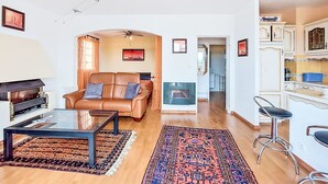 Uma sala de estar com sofá, mesa de centro e tapete, uma cozinha com armários e um banco de bar, e um corredor que leva a outro cômodo.