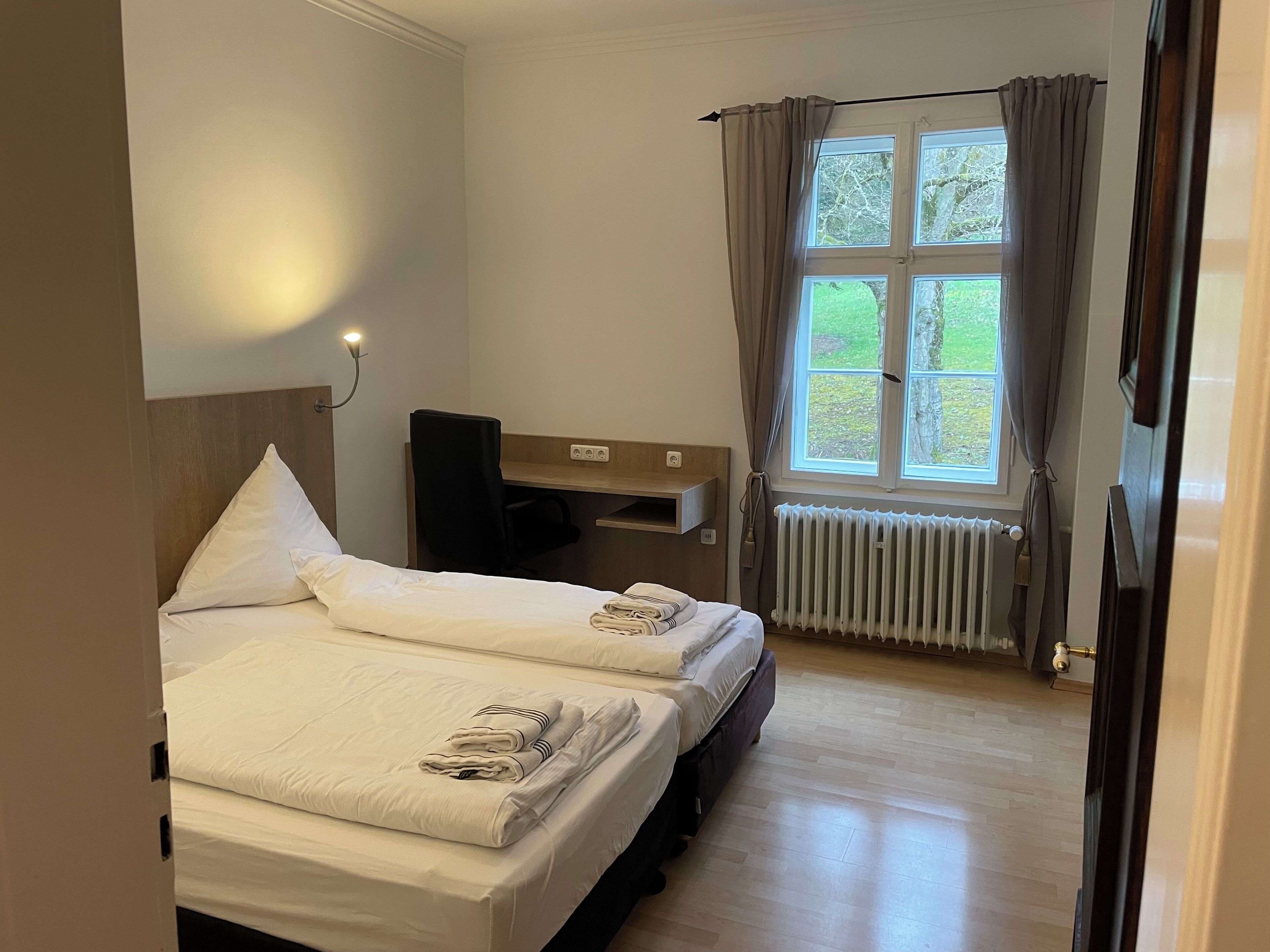 A hotel room with two beds, a desk, a chair, a window with curtains, and a radiator.