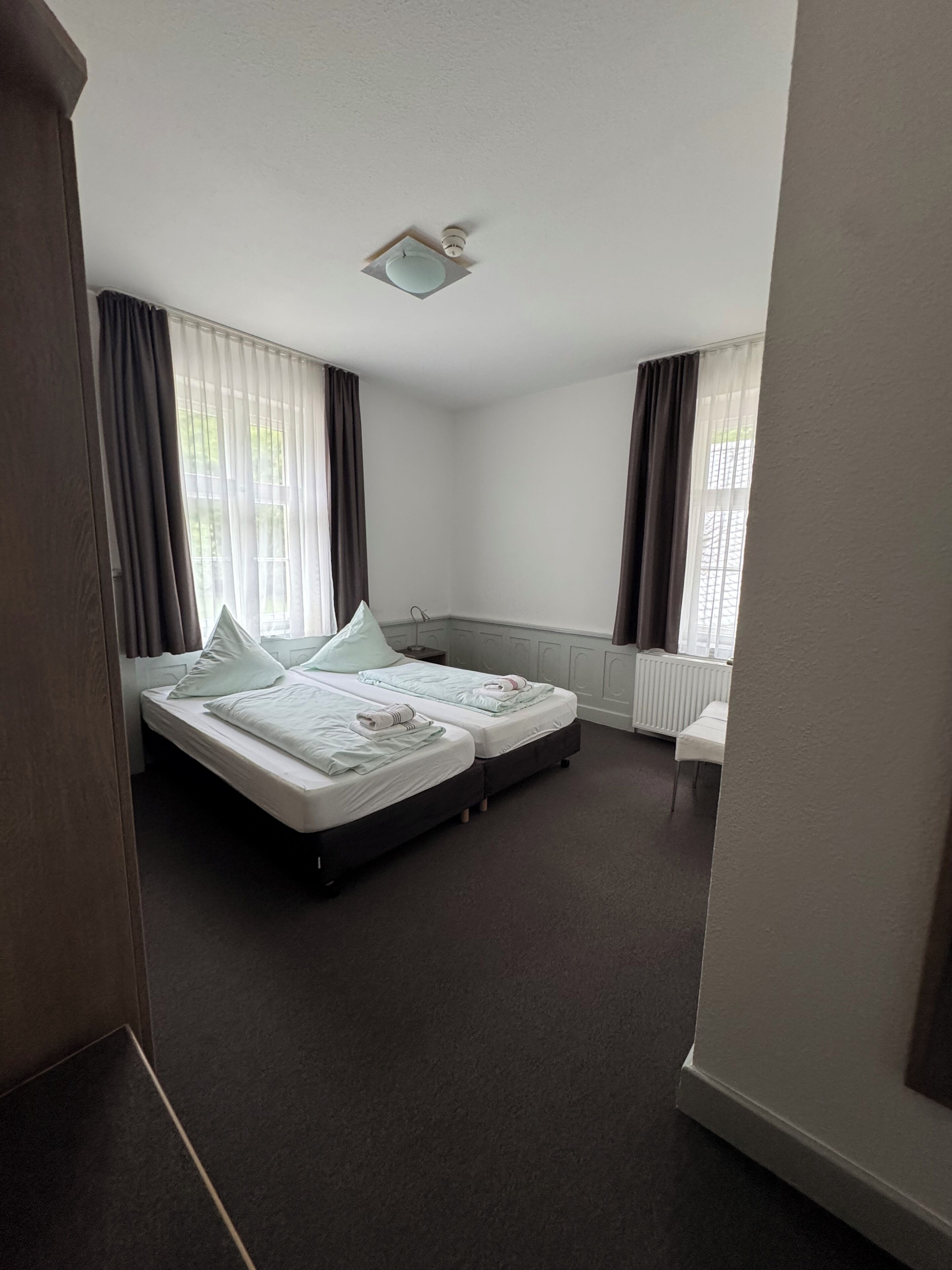 Habitación de hotel con cama doble, una mesita de noche pequeña, un radiador y una ventana con cortinas.