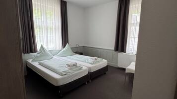 Habitación de hotel con cama doble, una mesita de noche pequeña, un radiador y una ventana con cortinas.