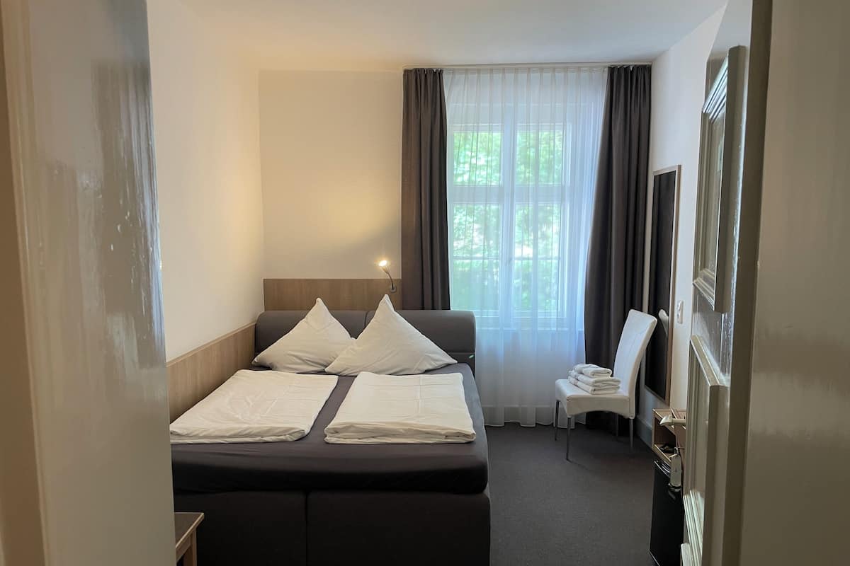 Una habitación de hotel con cama, una silla, un televisor y un ventanal con cortinas.