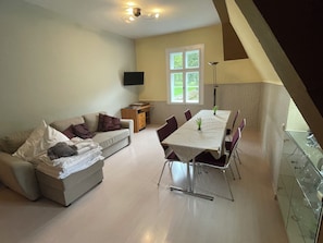 A living room with a sofa, a dining table with chairs, a TV mounted on the wall, and a window with curtains. - Landsitz Kammerburg - Verwalterwohnhaus (Lorch)