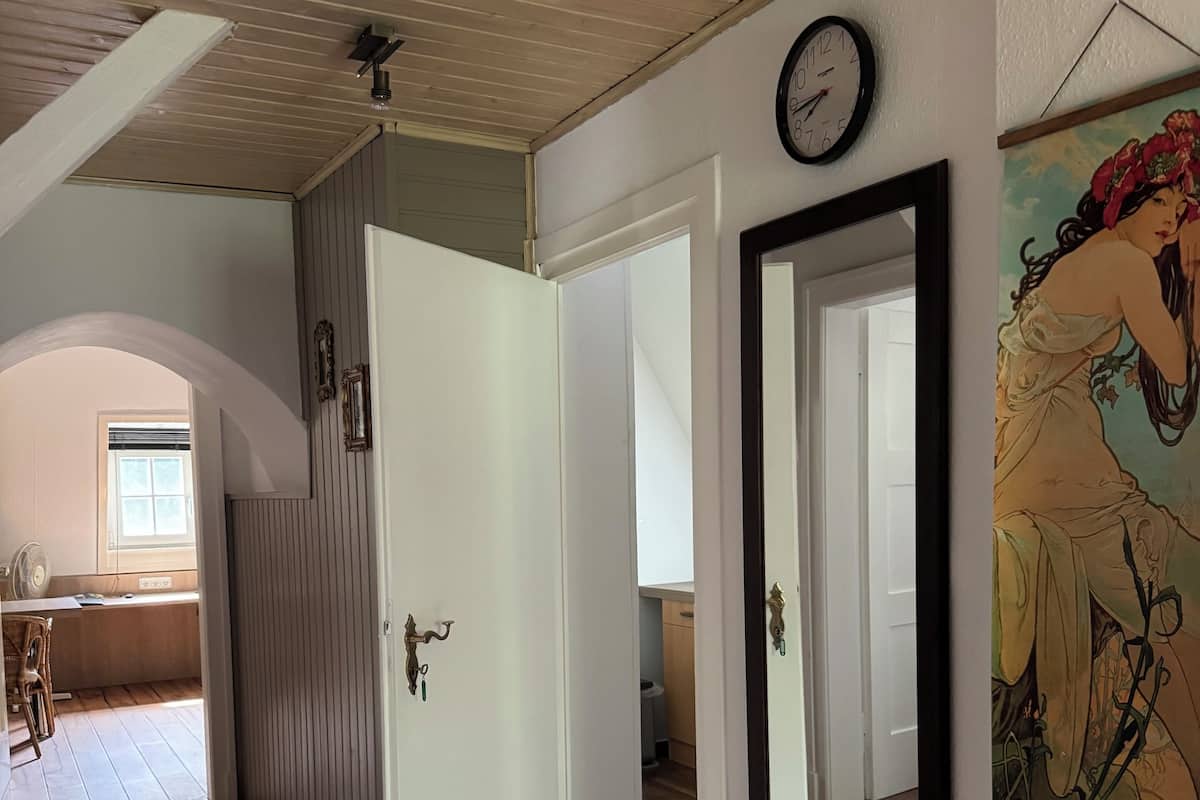 Un pasillo con suelo de madera, una puerta blanca, un reloj en la pared y una obra de arte enmarcada.