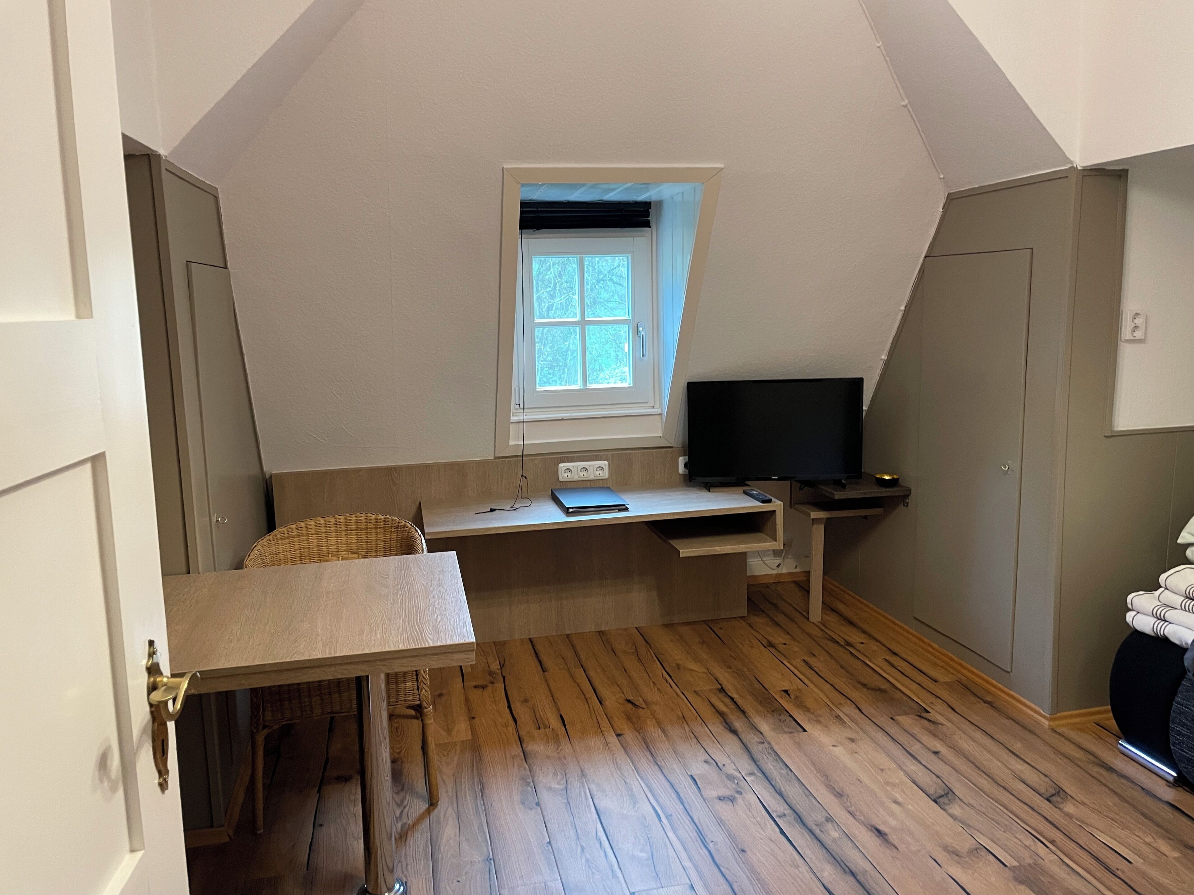 Een kamer met een schuine kap, een raam, een bureau met een computer, een stoel en een televisie.