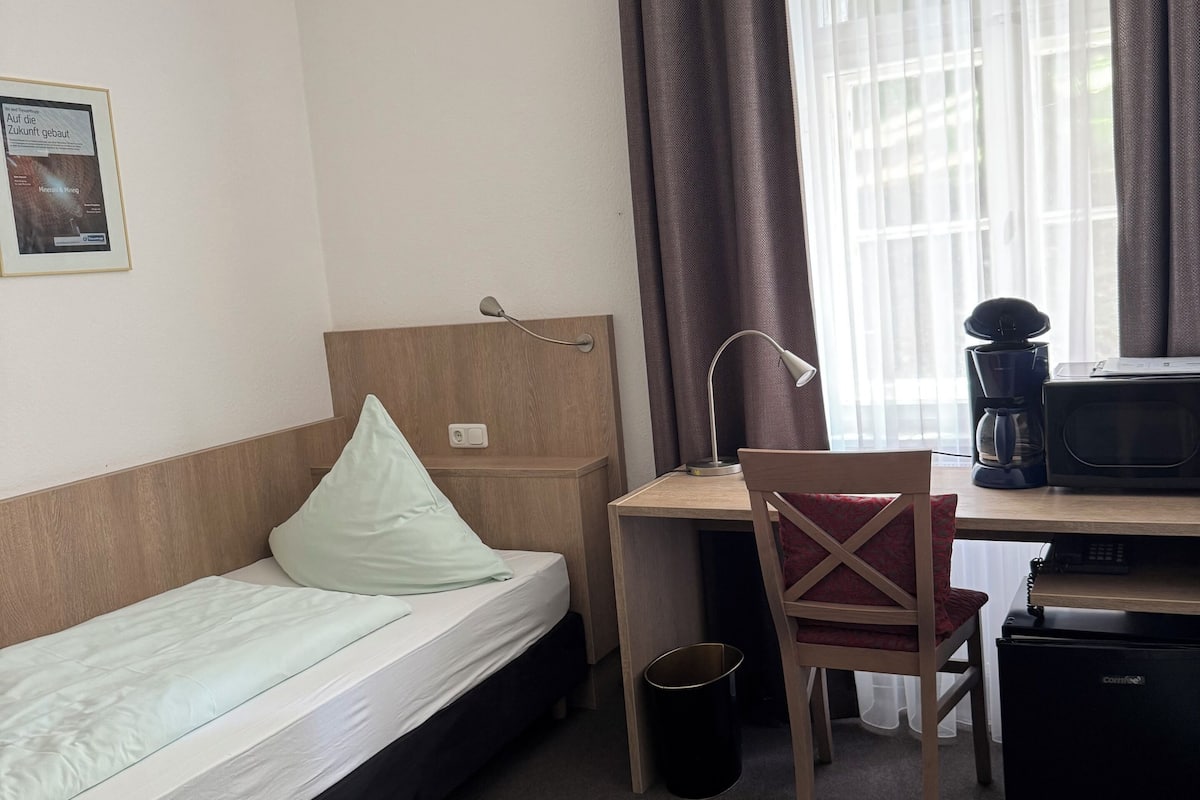 Habitación de hotel con una cama individual, un escritorio con silla, cafetera y un refrigerador pequeño.