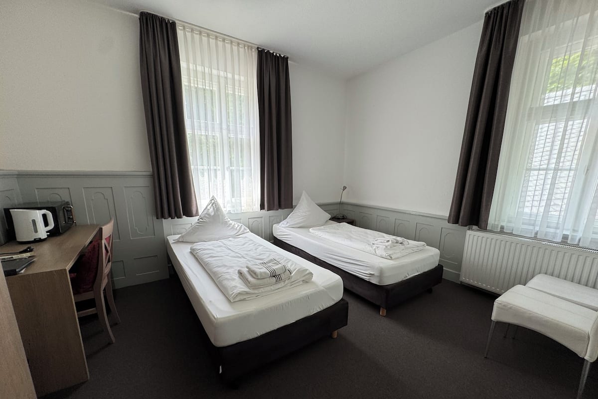 Habitación de hotel con dos camas individuales, un escritorio con cafetera, un radiador y ventanas amplias con cortinas.