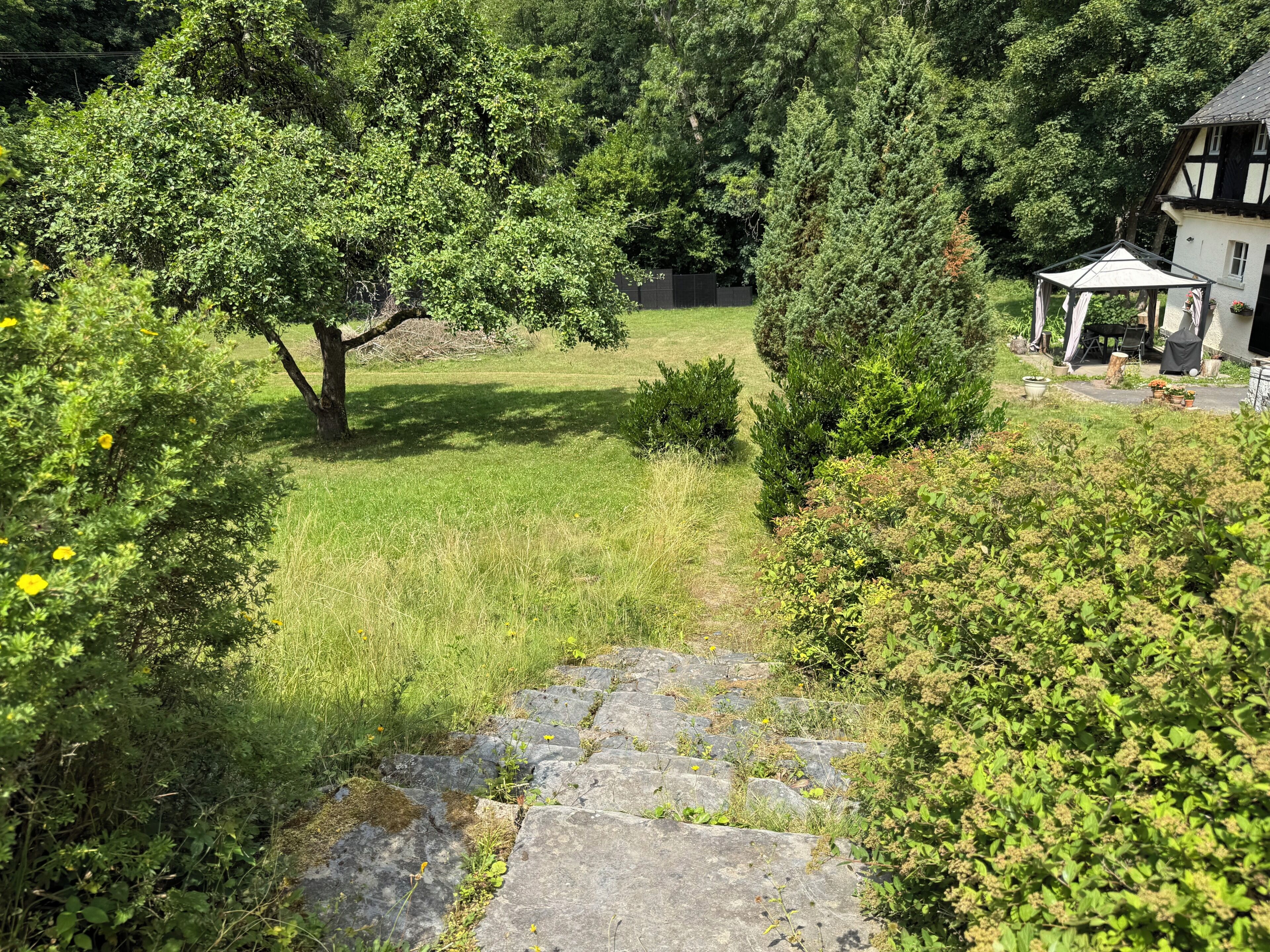 A stone pathway leading to a gazebo in a lush garden with a house in the background.