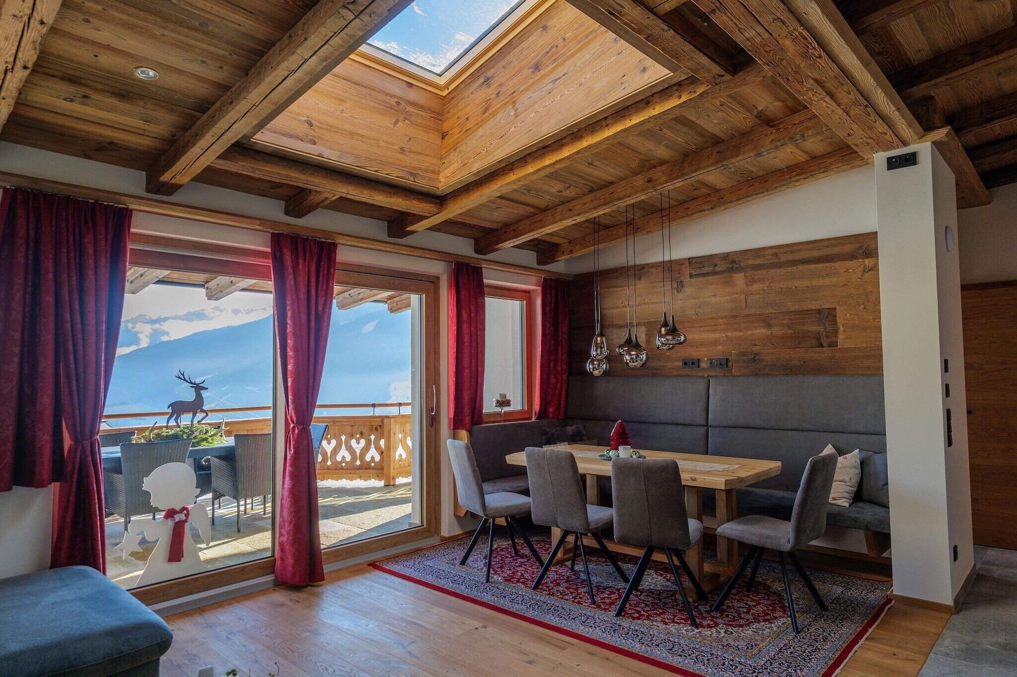 A room with a wooden ceiling, a dining table with chairs, a sofa, and a view of mountains through large windows.