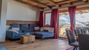 A living room with a sofa, coffee table, and chairs, a wooden ceiling, and a view of mountains through large windows. - Ski Apartment with panoramic views (Mittersill)