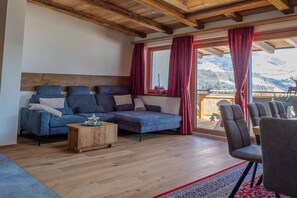 Ein Wohnzimmer mit einer Couch, einem Couchtisch und Sesseln, einer Holzbalkendecke und Bergblick durch große Fenster.