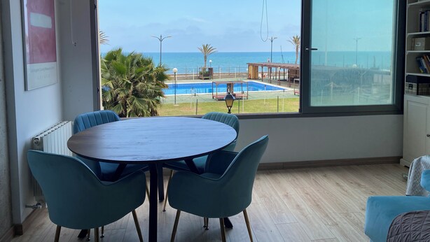 A room with a large window, a round table with chairs, a bookshelf, and a view of a pool and beach. - Port Saplaya Apartment (Alboraya)
