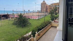 A balcony with a view of a beach, a tennis court, and a multi-story building. - Port Saplaya Apartment (Alboraya)