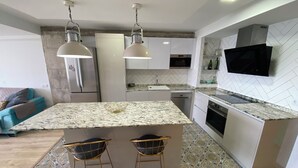 A modern kitchen with a marble countertop, two bar stools, a built-in island, stainless steel appliances, and a flat-screen TV mounted above the island. - Port Saplaya Apartment (Alboraya)