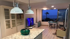 A modern kitchen with a marble countertop, two pendant lights, a television, and a view of the ocean at dusk. - Port Saplaya Apartment (Alboraya)