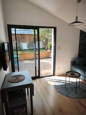 Uma sala de estar moderna com uma porta de vidro deslizante que dá para um deck de madeira, um sofá cinza, uma mesinha redonda e uma TV fixada na parede.