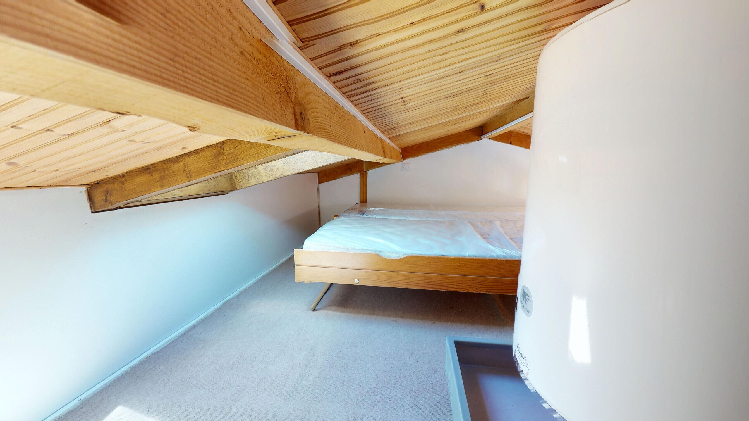 A small, narrow room with a single bed, wooden ceiling beams, and a white wall.
