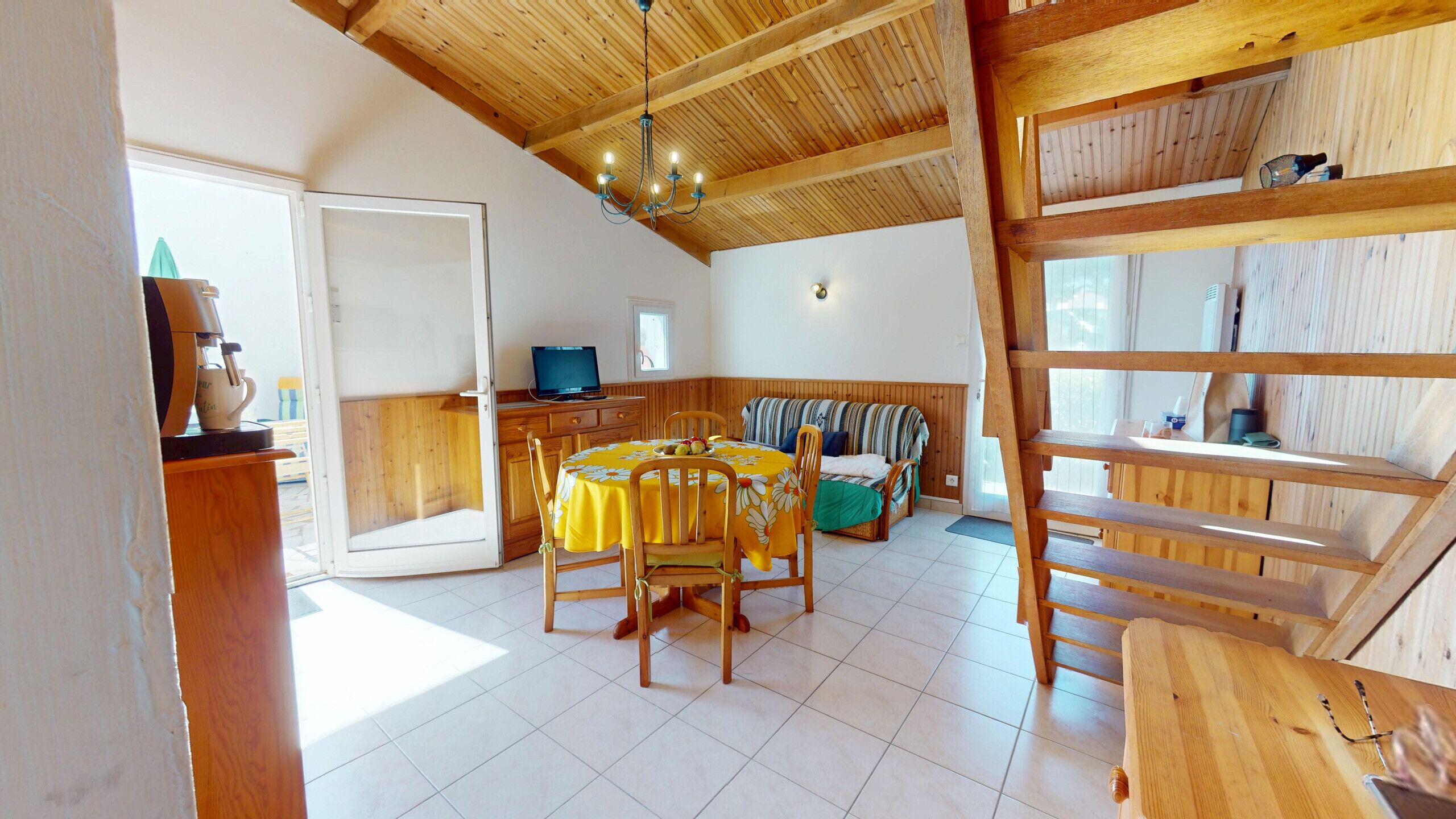 Ein gemütliches Zimmer mit Holzbalkendecke, einem Esstisch mit Stühlen, einer Couch, einem Fernseher und einer Treppe.