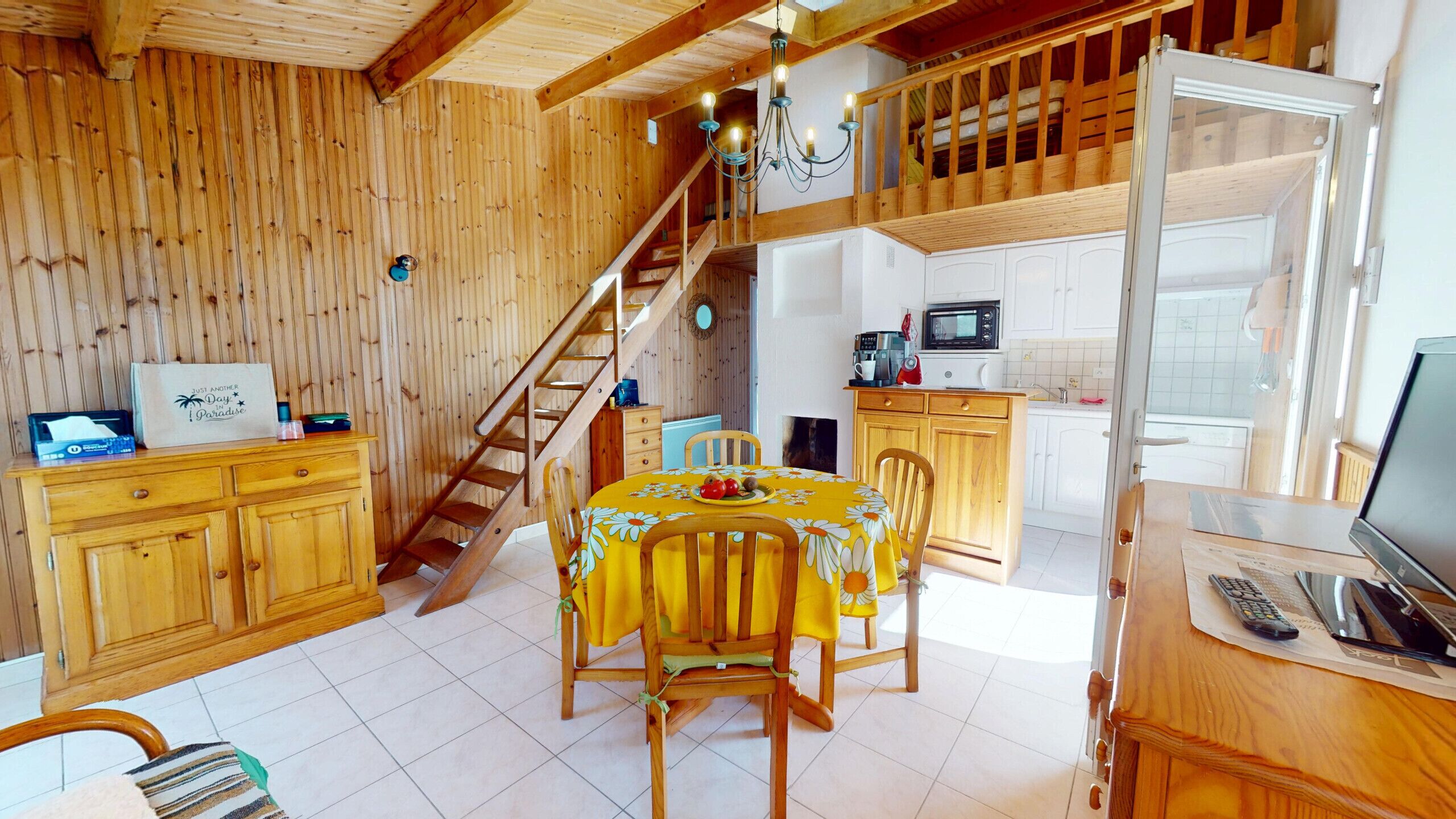 A wooden interior with a dining table, chairs, a staircase, kitchen area, and a TV.