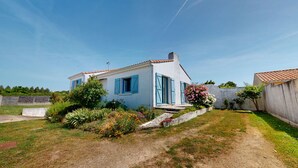 A two-story house with blue shutters, a garden with various plants and flowers, and a clear blue sky. - Villa Good Vibes - Bikes available, large fenced-in garden (Le Fenouiller)
