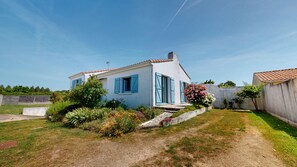 Ein zweistöckiges Haus mit blauen Fensterläden, ein Garten mit verschiedenen Pflanzen und Blumen, und ein klarer, blauer Himmel.