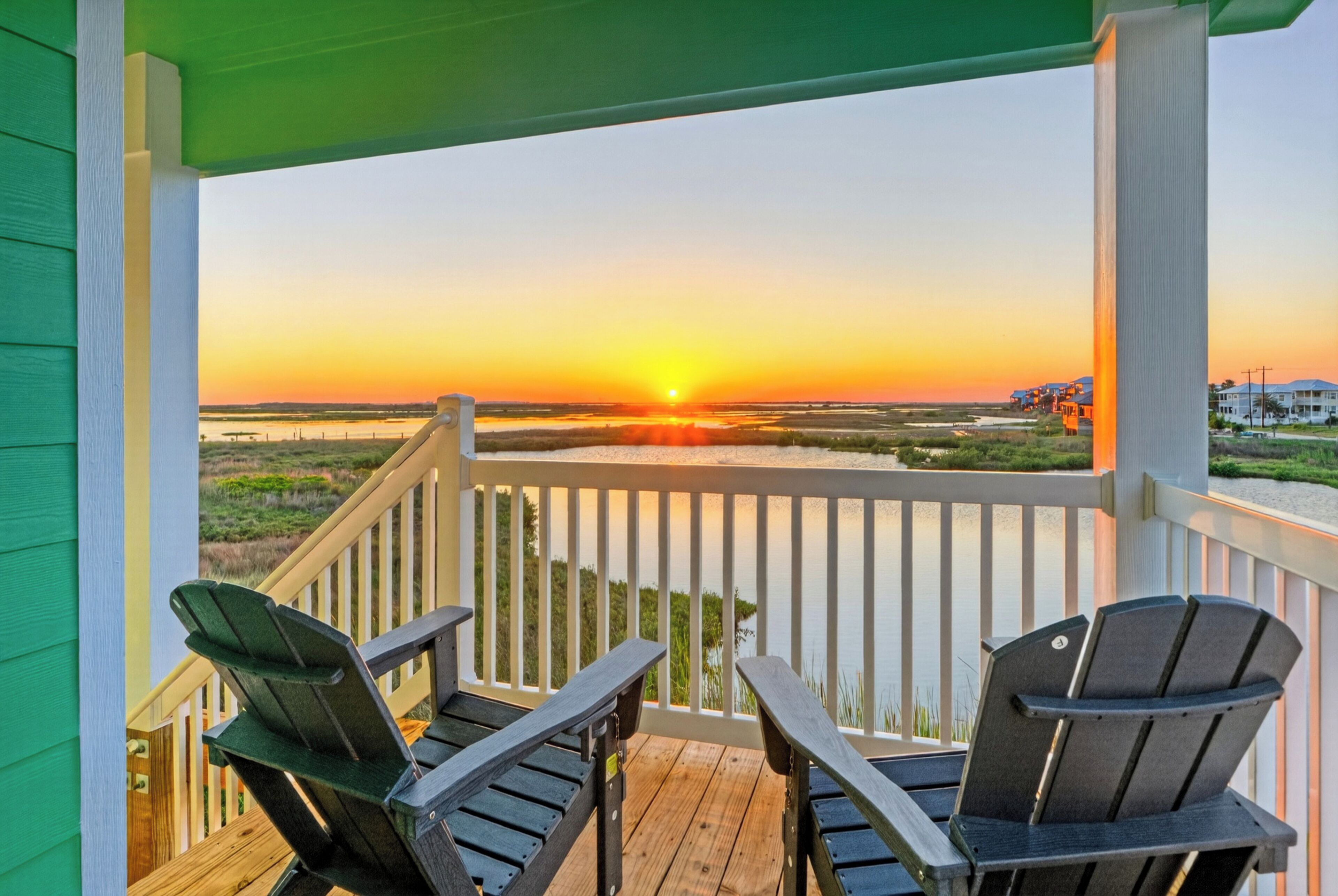 A wooden deck with two Adirondack chairs facing a sunset over a water body.