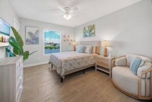 Ein Schlafzimmer mit Bett, Stuhl, Nachttisch, Deckenventilator und Fenster mit Jalousien.
