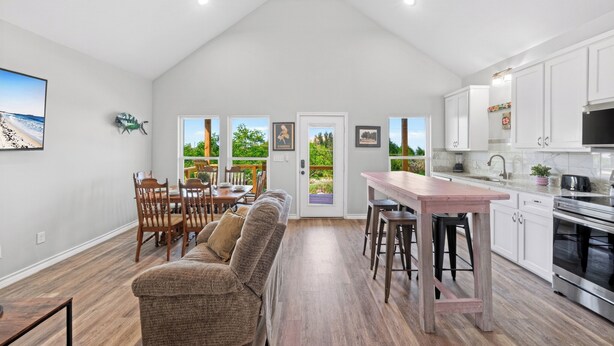 A modern kitchen with white cabinets, a dining area with a wooden table and chairs, and a view through a glass door to an outdoor patio. - Water Views | Wraparound Decks | Pool Access | Flour Bluff Bay | Black Drum (Corpus Christi)