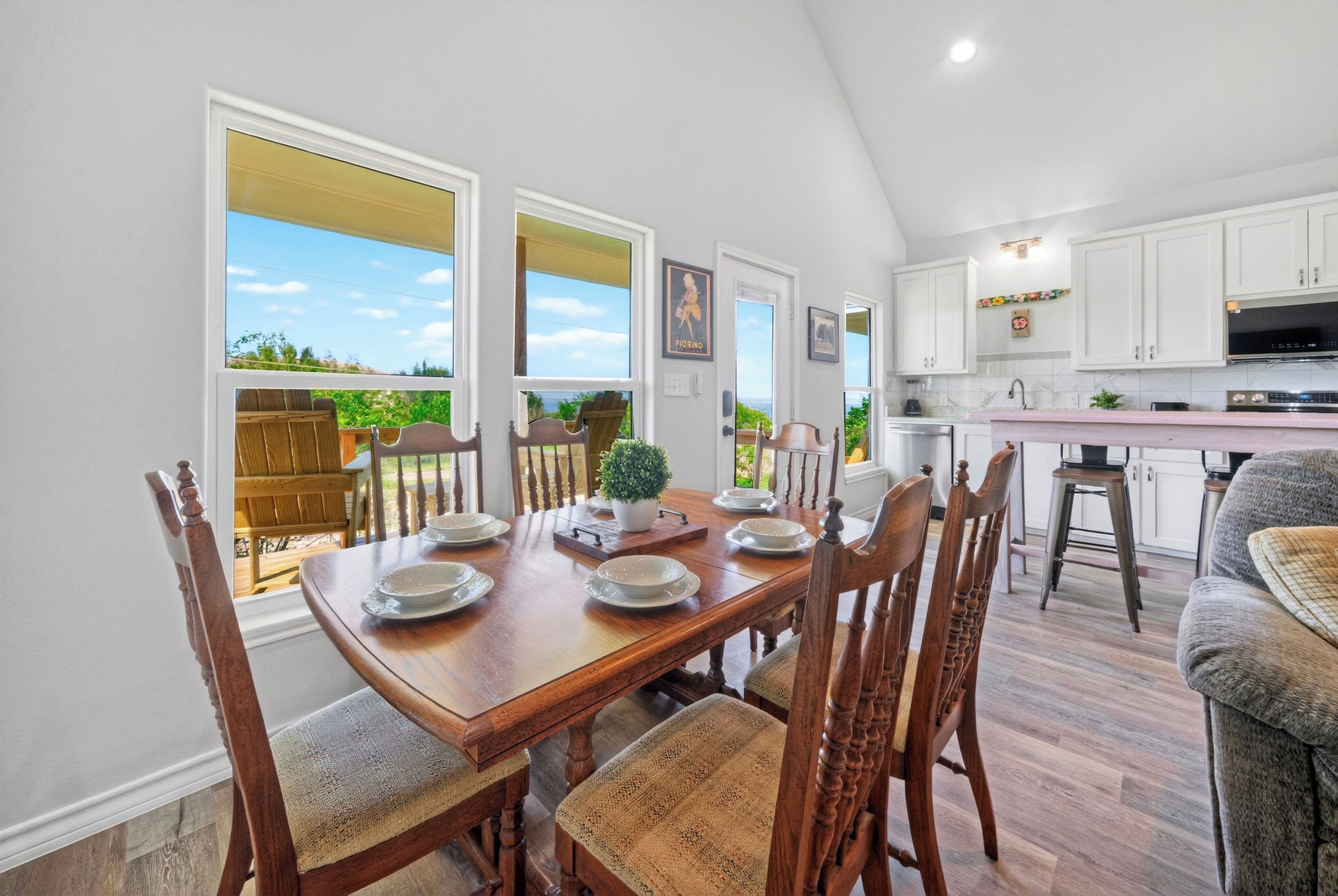 A dining area with a wooden table set for four, surrounded by chairs, with a view of the outdoors through large windows.