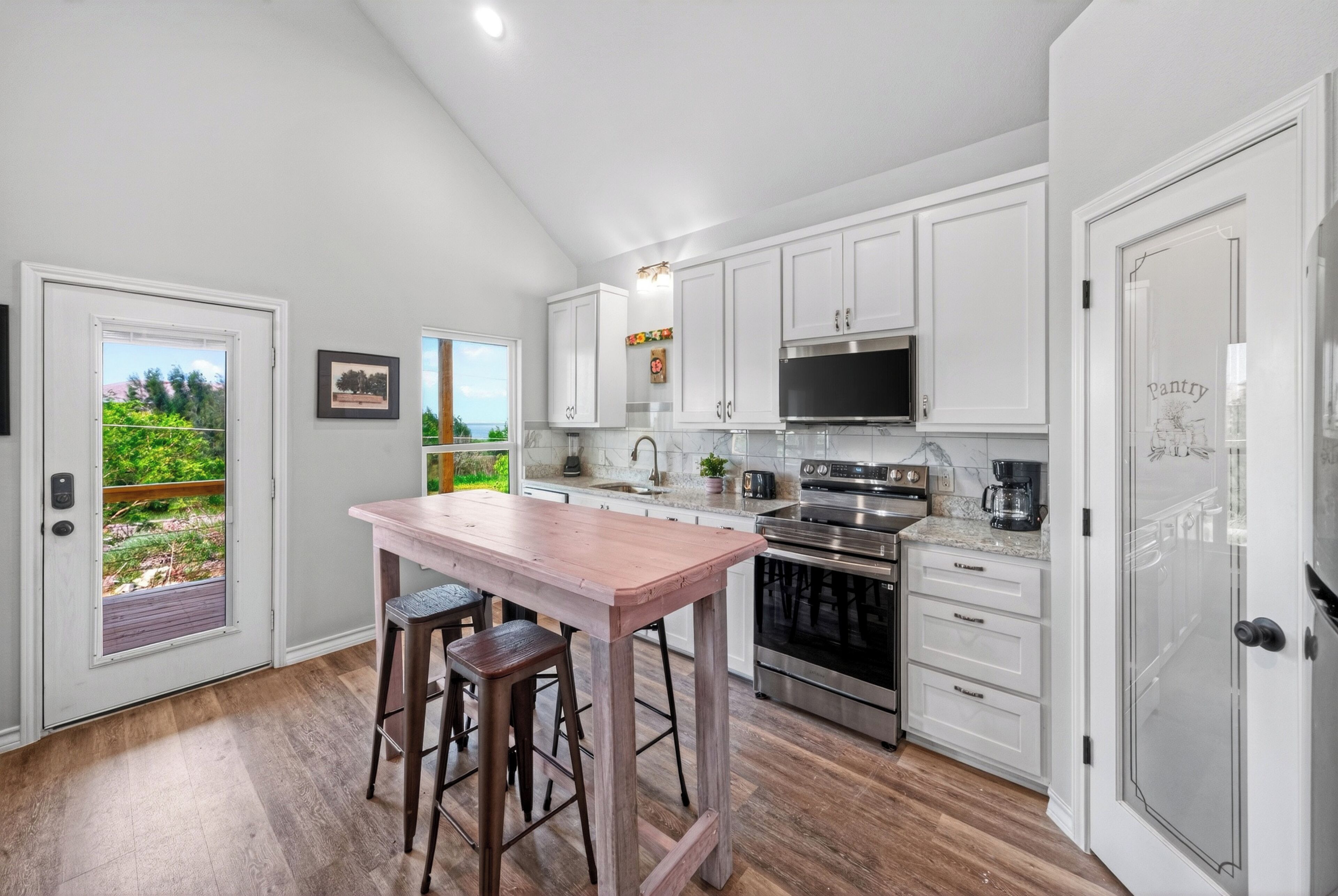 A kitchen with white cabinets, a wooden island with stools, a stainless steel oven, and a view through a door to an outdoor area.