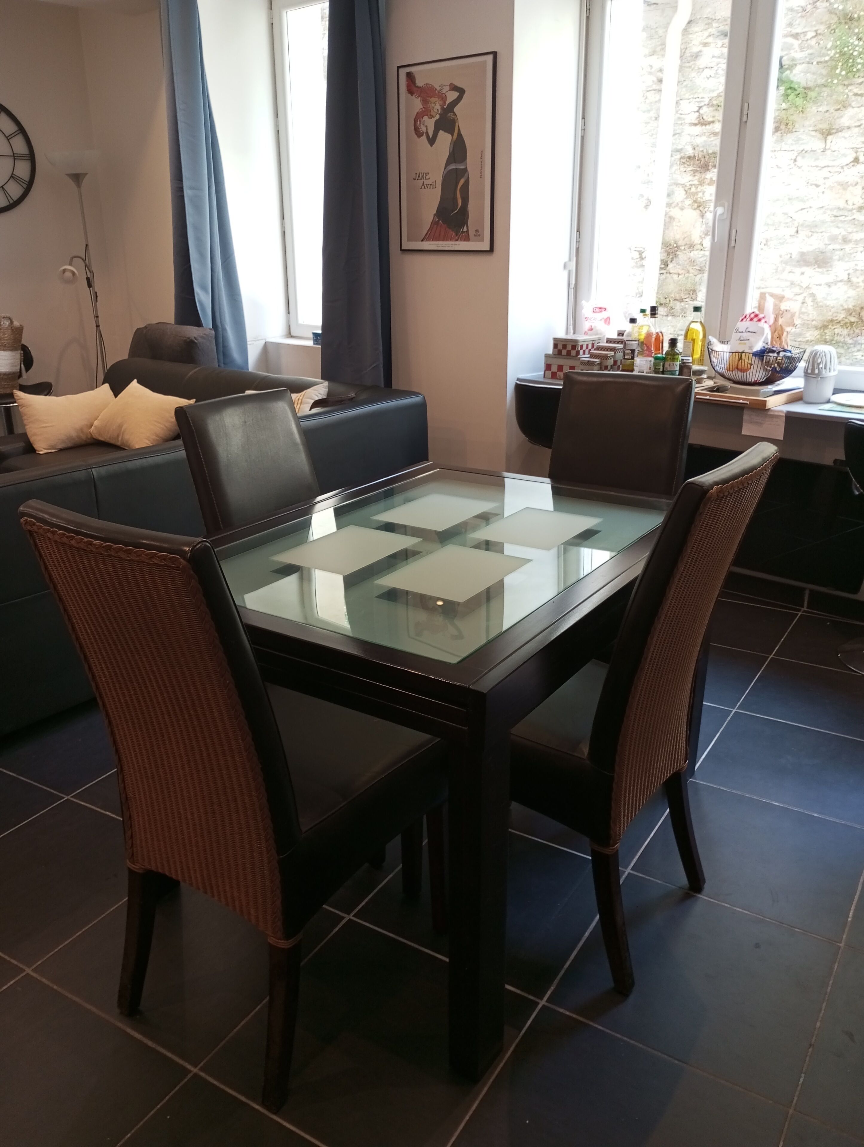 Een eethoek met een tafel met glazen blad, donkere stoelen, een bank en een wandklok.