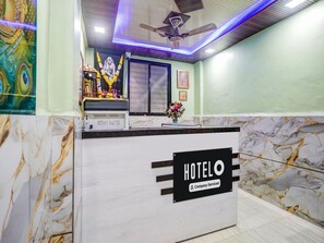 A hotel reception desk with a 'HOTELO Company-Serviced' sign, a ceiling fan, and a decorative garland.