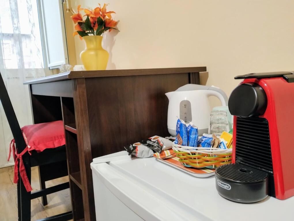 Een hotelkamer met een houten bureau, een witte waterkoker, een rode koffiezetapparaat, een mand met snacks en een vaas met bloemen.