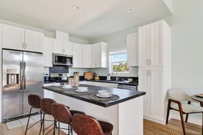 A modern kitchen with white cabinets, a stainless steel refrigerator, a microwave, and a large island with bar stools. - Sun-Filled Two-Story Home | Near Balboa Park + Gaslamp Quarter | J Street (San Diego)
