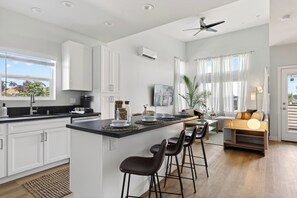 A modern kitchen with white cabinets, a central island with bar stools, a ceiling fan, and a dining area with a sofa and lamp. - Sun-Filled Two-Story Home | Near Balboa Park + Gaslamp Quarter | J Street (San Diego)