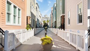 A wooden boardwalk with white railings, lined with colorful buildings and a large planter with yellow foliage. - Steps to the Beach + Resort Pools + Golf | Wild Dunes IOP | 137 Grand Pavilion (Isle Of Palms)