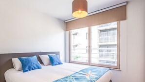 A bedroom with a bed, a window with a view of buildings, a hanging light fixture, and a starfish design on the bedspread. - Chateau Nouvella with balcony by Interhome (Blankenberge)