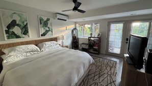 A bedroom with a bed, ceiling fan, artwork, air conditioning unit, and a television.
