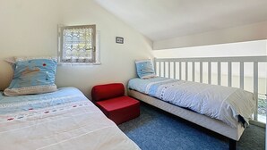 Quarto com duas camas, uma cadeira vermelha, janela com cortinas estampadas e corrimão branco.