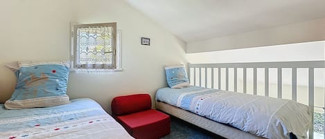 Une chambre avec deux lits, un fauteuil rouge, une fenêtre avec des rideaux à motifs et une rambarde blanche.