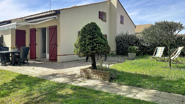 Um edifício térreo com telhado de telhas, uma porta com persiana vermelha e uma pequena área de jardim com um arbusto aparado e uma cadeira.