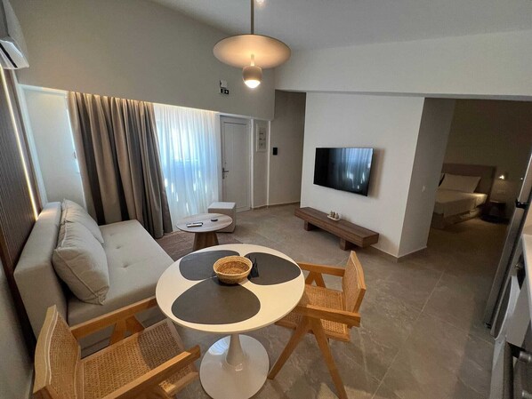A modern living room with a sofa, round dining table, wicker chairs, a TV, and a bench. - Ixia Cozyline Apartments - 105 Studio (Rhodes)