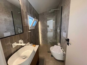 A modern bathroom with a large sink, a toilet, a glass shower enclosure, and a mounted television. - Ixia Cozyline Apartment- 103 (Rhodes)