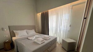 A modern hotel room with a large bed, a bedside table, a small stool, and a window with sheer curtains. - Ixia Cozyline Apartment - 104 (Rhodes)